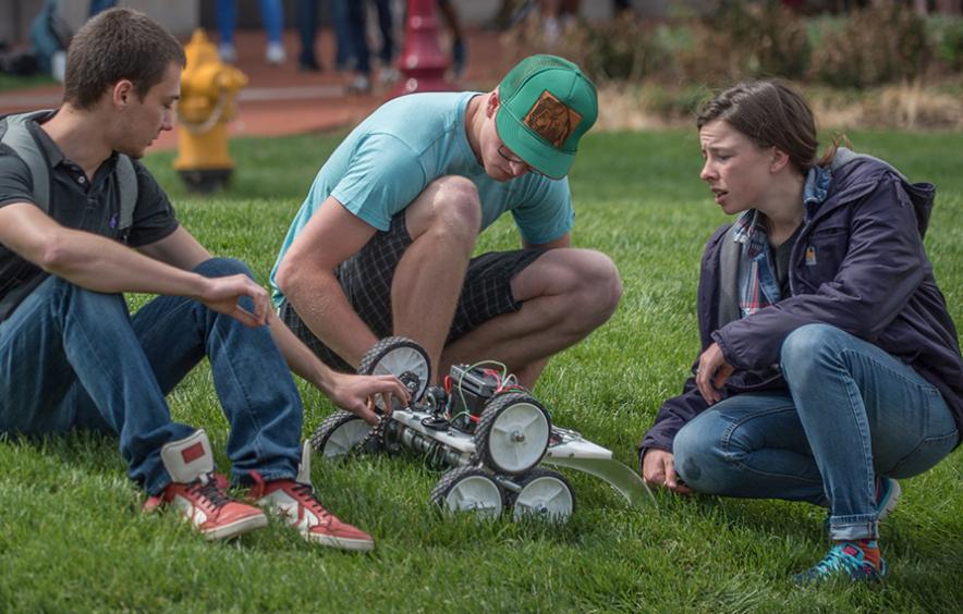Students gather on the lawn to discuss a robot they're constructing.